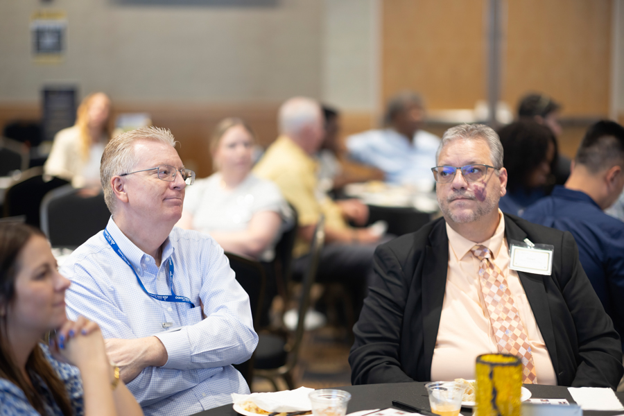 People attend a conference or seminar, with two men in the foreground: one in a light blue shirt with a lanyard and the other in a suit with a name badge at a round-table gathering.