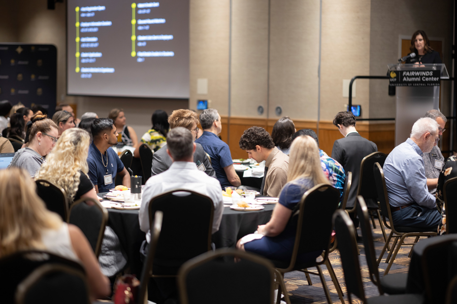 Audience seated at round tables in a banquet-style room, listening to a speaker at a podium on a stage.