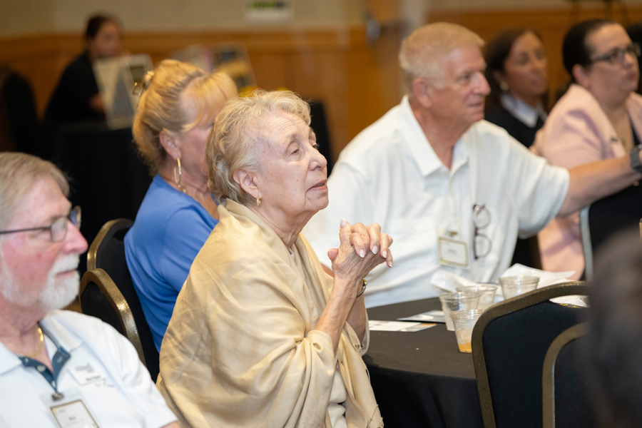 Elderly woman in a beige shawl sits at a round table, hands clasped, attentively listening at a conference.