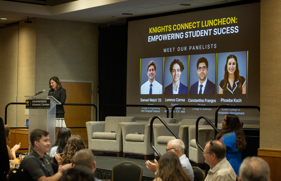 Speaker at podium on a stage during the Knights Connect Luncheon; a large screen behind shows four panelists with names.