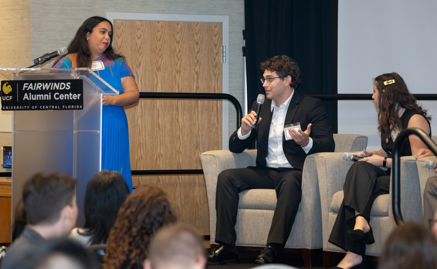 Woman in a blue dress stands at a podium labeled Fairwinds Alumni Center as a man in a suit and a woman in black sit onstage and speak into microphones, with audience in the foreground.