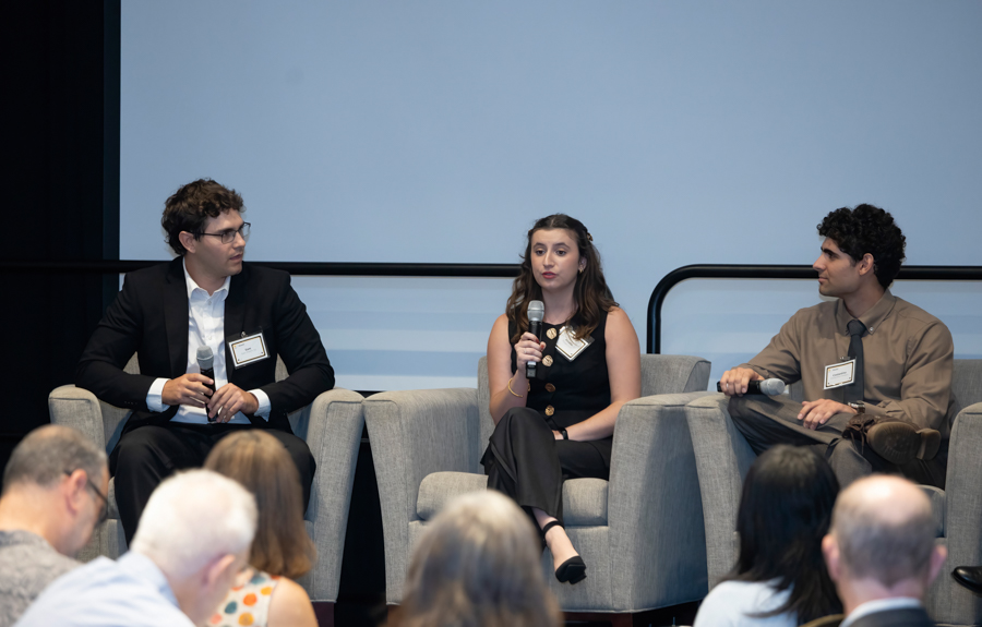 Three panelists sit on stage in gray chairs, each holding a microphone during a panel discussion with an audience in front of them.