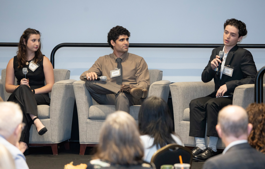 Three panelists seated on stage in gray chairs, each with a microphone; audience visible in foreground.