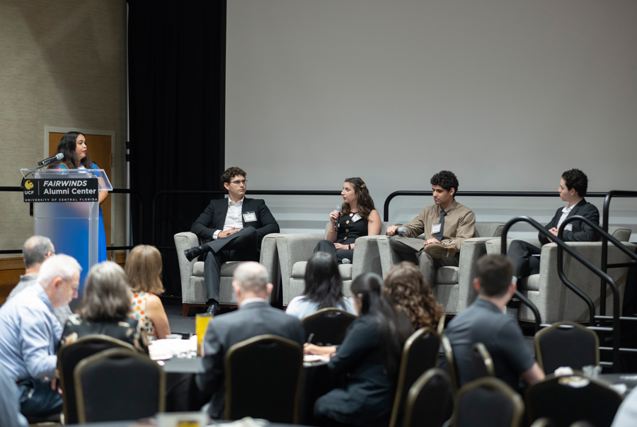 Panel discussion on stage with a speaker at a podium and four panelists, audience seated at round tables in front. The podium branding reads 'Fairwinds Alumni Center.'