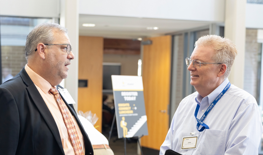 Two professionally dressed men with name badges chatting in a bright conference lobby.