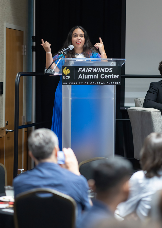 Woman in a blue dress speaks at a lectern labeled 'Fairwinds Alumni Center, University of Central Florida' with a microphone.
