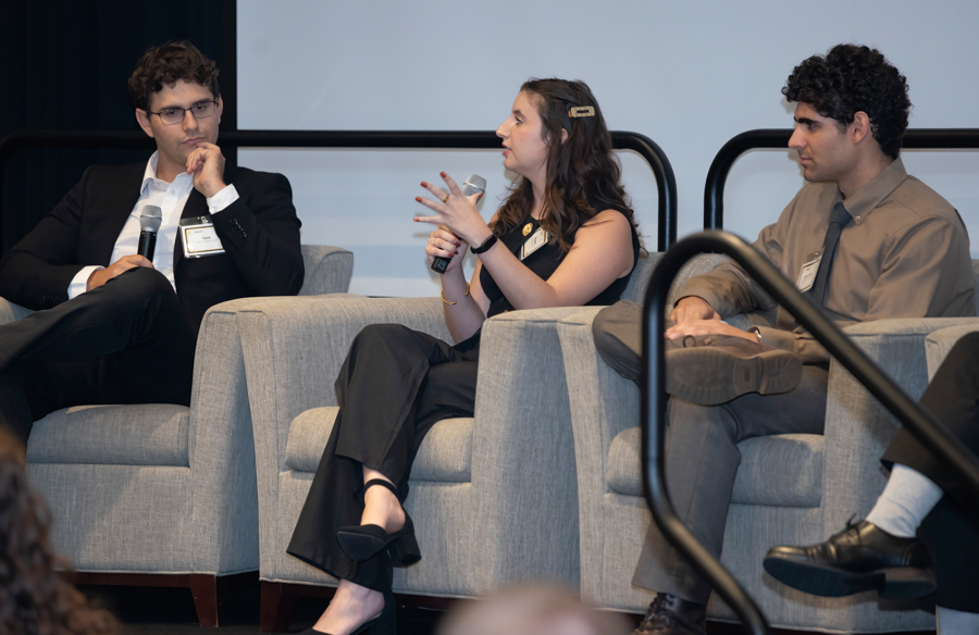 Three panelists seated on a gray couch on stage; the woman center speaks into a microphone while the men listen nearby.