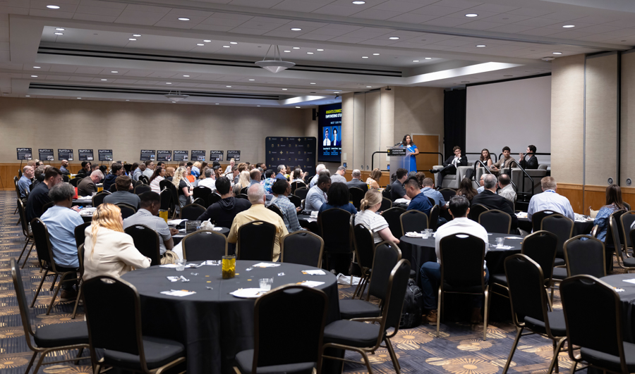 Speaker at a podium presenting to a large audience in a conference room or hotel ballroom.