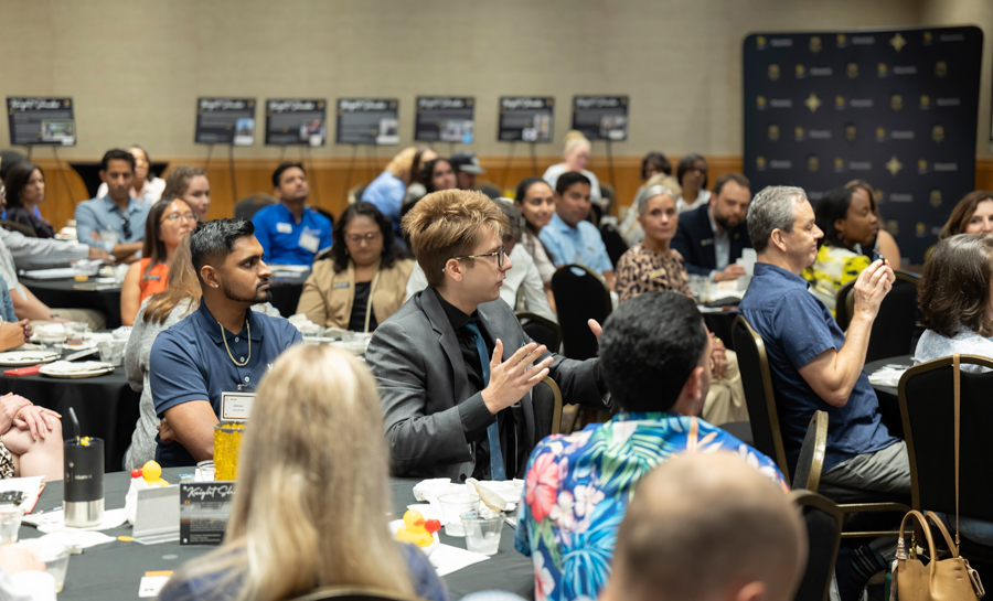 Man in a gray suit gestures while speaking at a round-table conference with diverse attendees listening.