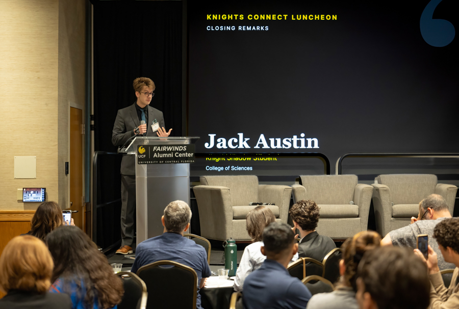 Speaker at a podium presenting to a room full of attendees, with a large backdrop reading 'Jack Austin' and 'Knights Connect Luncheon' on stage at Fairwinds Alumni Center in the background.