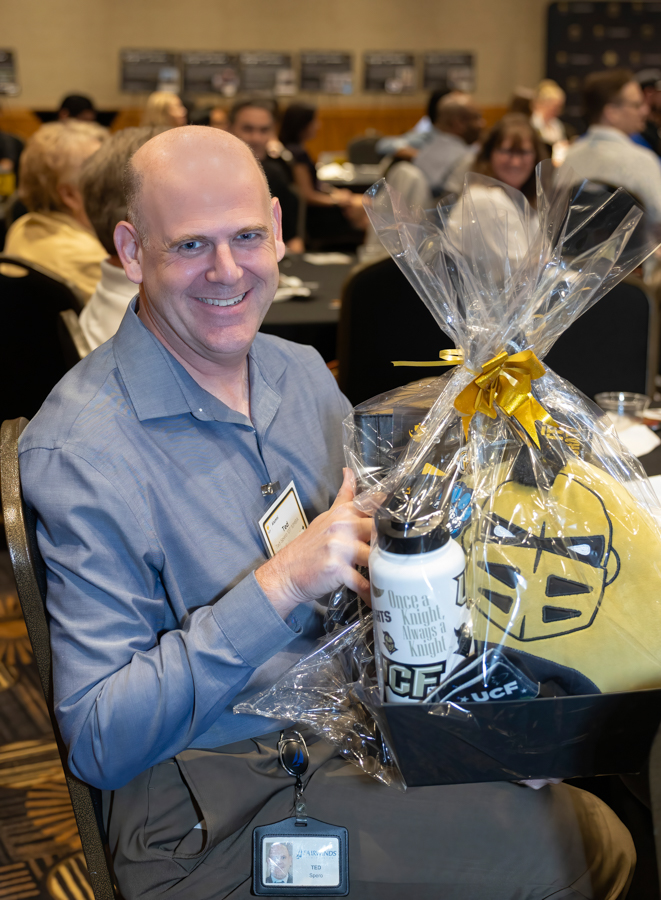 Smiling man in a blue shirt seated at a conference table, holding a gift basket wrapped in clear cellophane with a yellow ribbon, containing a mug and a yellow plush item.