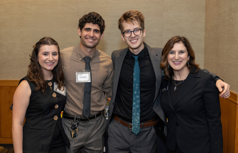 Four adults stand together, smiling for a group photo at a formal event, arms around each other.