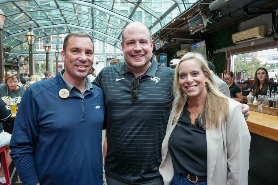 Three smiling adults posing together at a busy indoor bar with a glass roof and warm lighting.