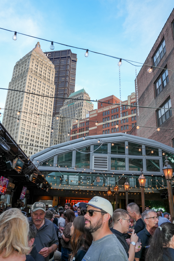 Crowded outdoor city venue beneath string lights, with a glass-roofed pavilion and tall skyscrapers in the background.