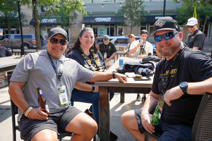 Three friends sit at an outdoor table on a sunny street, smiling and holding beers with lanyards around their necks. They chat at a casual outdoor gathering near storefronts and parked cars.