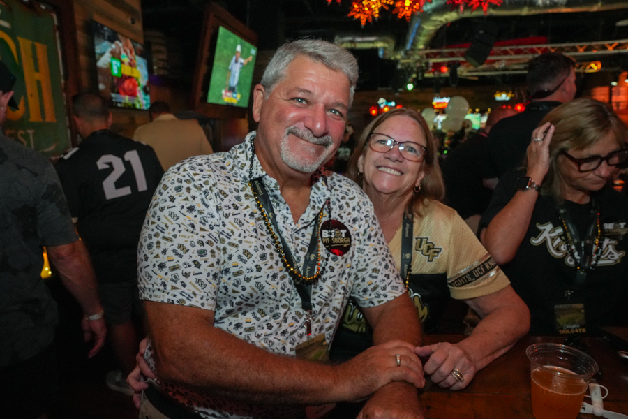 Smiling couple posing at a crowded bar, man in a patterned shirt with a badge and beads, woman beside him with glasses and lanyard, drinks on the counter behind them.