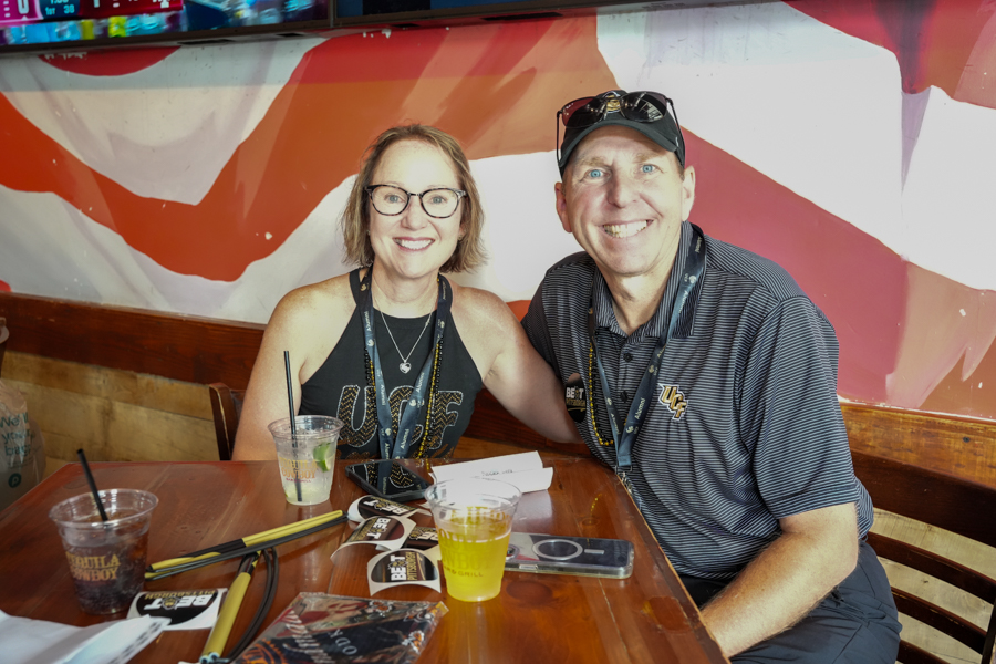 Two smiling adults sit at a wooden table with drinks in front, against a colorful mural backdrop.