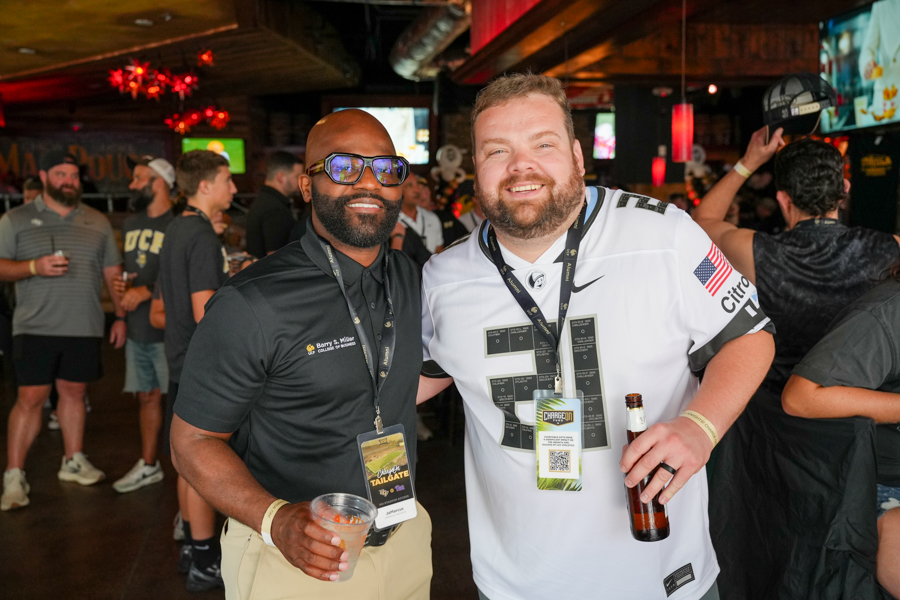 Two smiling men pose for a photo at a crowded bar/event; one wears a black polo and sunglasses, the other a white jersey, both with lanyards and drinks nearby.