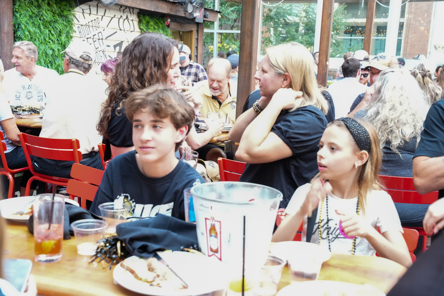A young boy and girl sit at a wooden outdoor cafe table with drinks and plates, while a crowd chats at surrounding tables in the background.