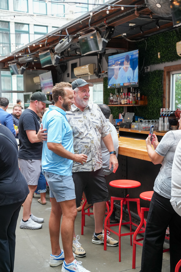 Two men smile and pose together at a busy bar, one in a blue polo and the other in a patterned shirt, holding drinks.