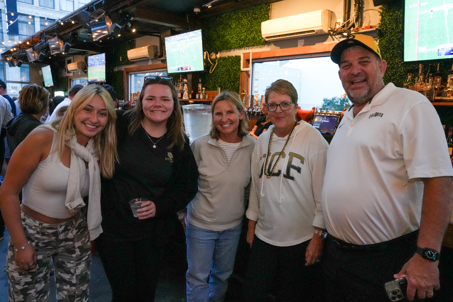 Five smiling adults pose together at a bar, with multiple TV screens showing a sports game in the background.
