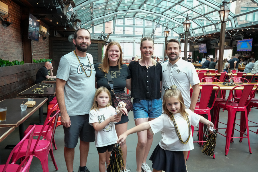 Six people pose for a group photo in a bright indoor bar/restaurant with red chairs and string lights, two young girls in front holding pom-poms.