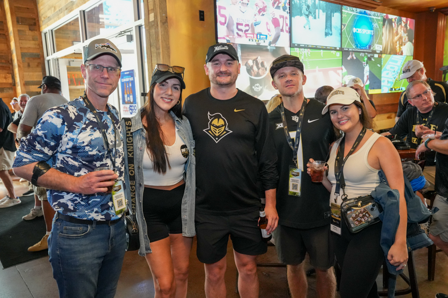 Five people pose together at an event space with multiple TVs behind them, wearing lanyards and holding drinks.