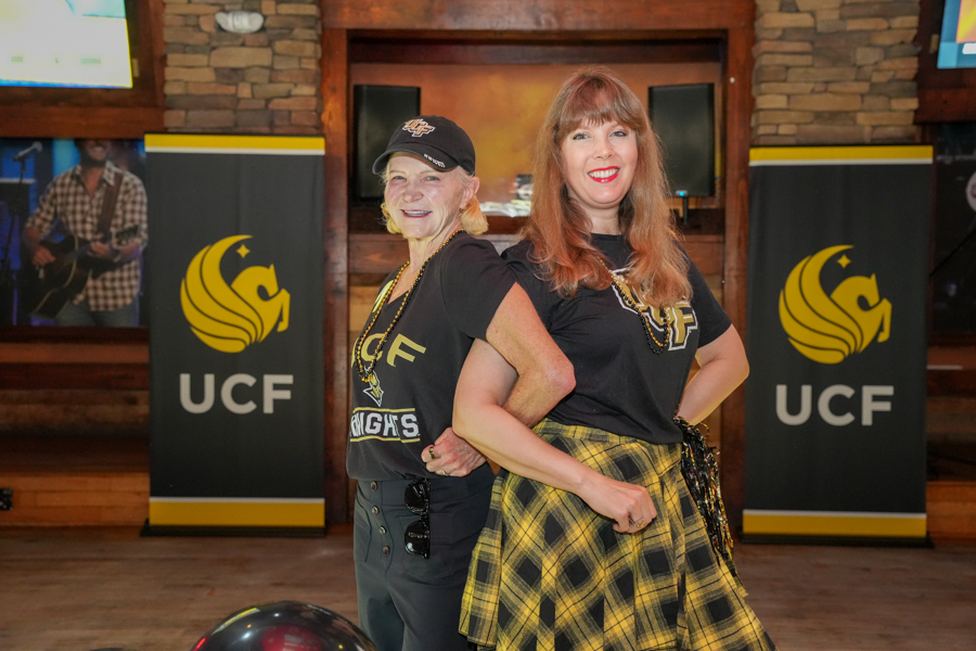 Two women stand back to back in a bar or event venue, both wearing black UCF-themed shirts and smiling at the camera, with UCF banners in the background.