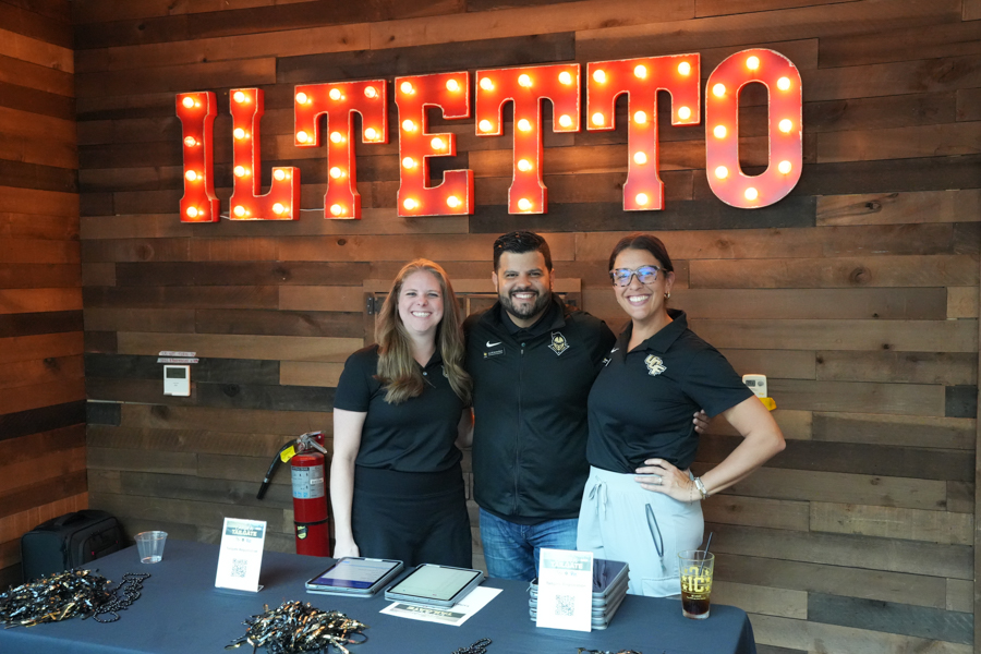 Three people stand behind a booth table with a lit ILTETTO sign in the background.