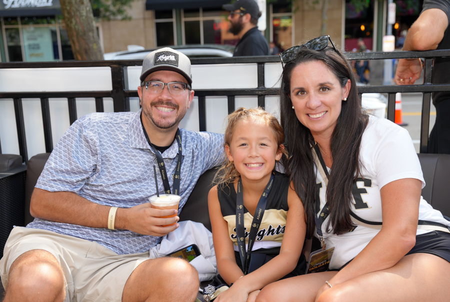 Smiling family of three sitting on outdoor seating at an event: man with cap and drink, girl in the middle, woman on the right wearing a white shirt.
