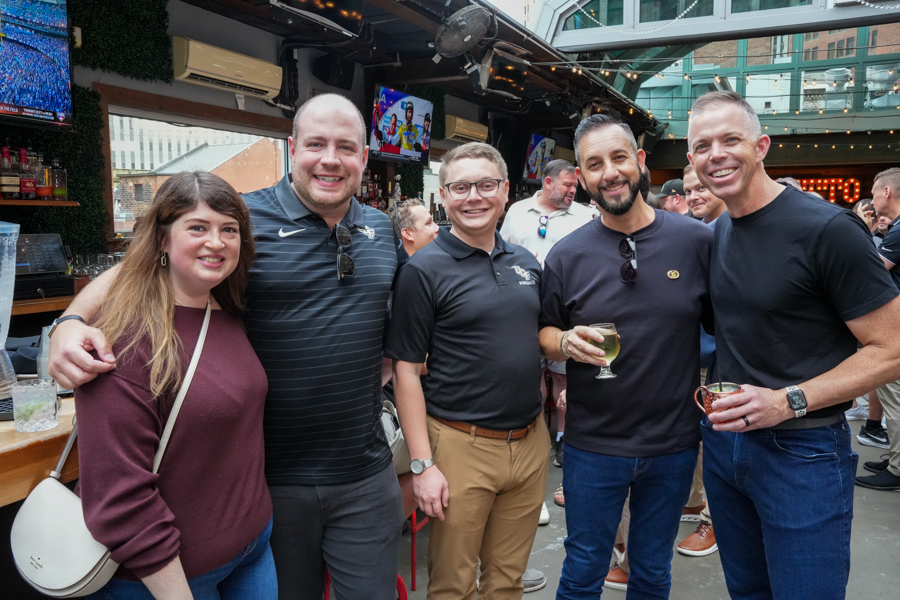 Five friends posing together at a bar patio, smiles and drinks in hand with a lively crowd behind them.