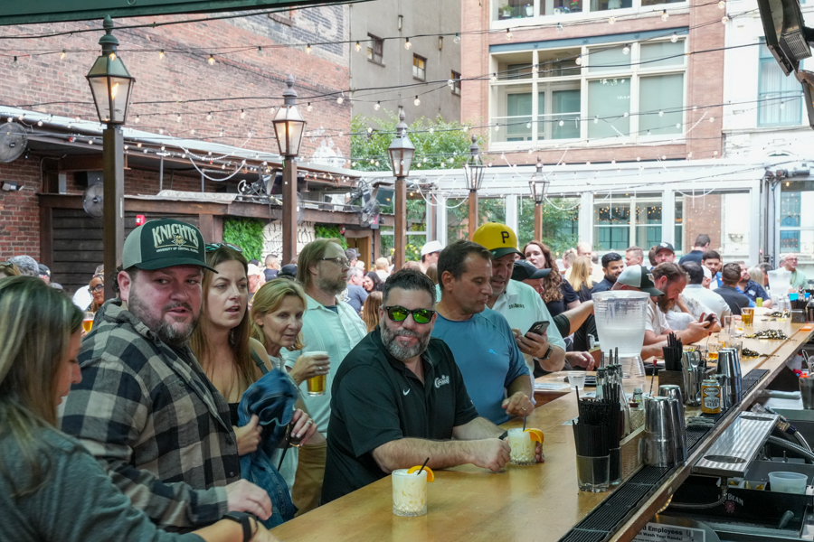 Crowded outdoor bar scene with patrons at a long wooden counter under string lights and brick buildings in the background.