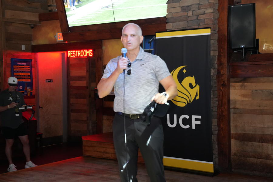 Male speaker on stage holding a microphone, with a UCF banner and wood-paneled backdrop in a casual venue.