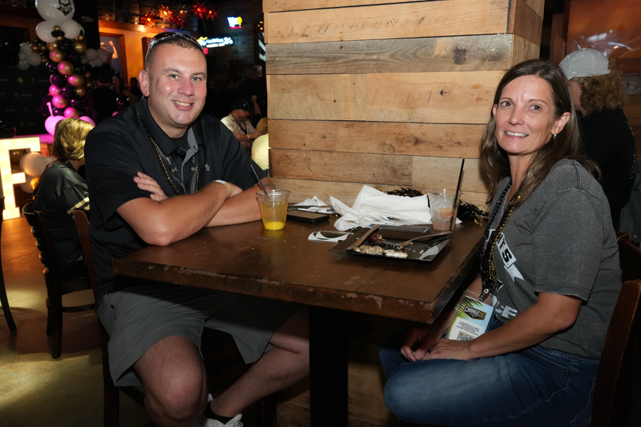 Two adults sit at a wooden table in a lively restaurant, smiling at the camera with drinks and a plate between them.