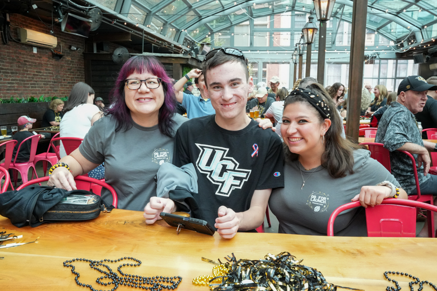 Three friends sit at a wooden table in a bright, glass-walled venue, smiling at the camera; two wear gray 'Go For Launch' shirts and pink/black beads on the table nearby.09