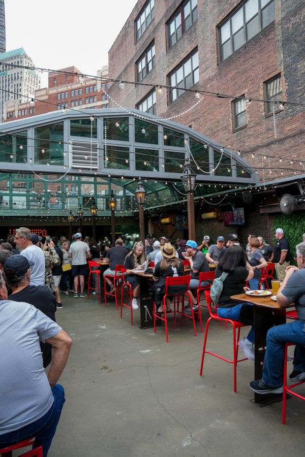 Outdoor urban courtyard restaurant with string lights, red metal chairs, and diners under a glass canopy attached to brick buildings.] , should be concise but we must remove stray quotes.