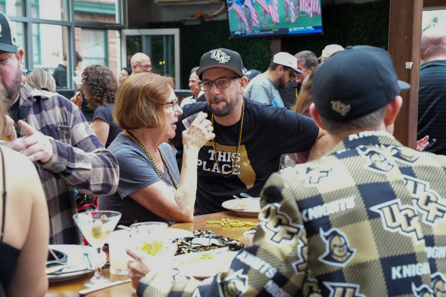 Woman with glasses talks to a man in a black cap at a crowded outdoor gathering, drinks on the table.