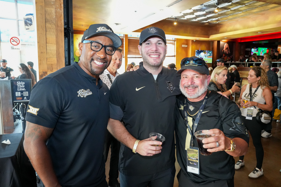 Three men pose for a photo at a busy indoor event, smiling and holding drinks, wearing dark polo/shirt logos.