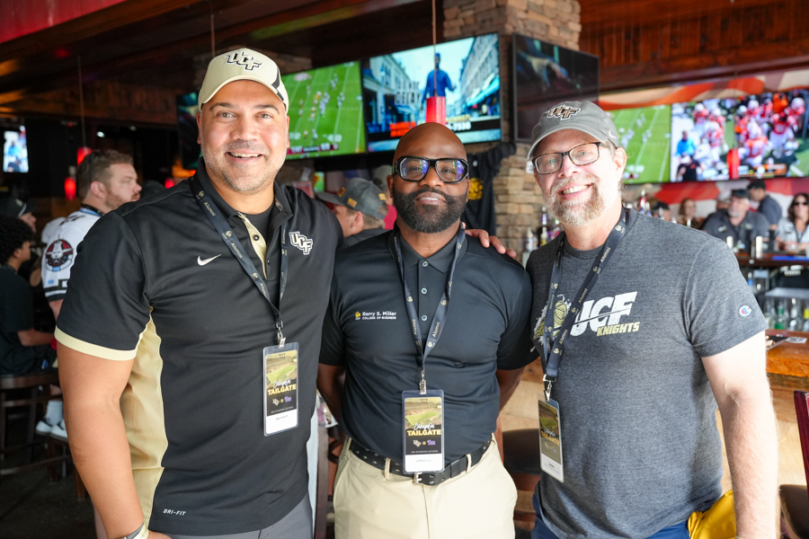 Three men posing together at a sports bar tailgate, wearing UCF apparel and lanyards.