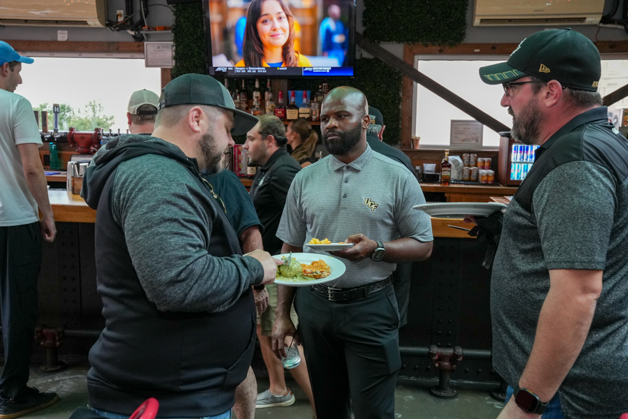 Three men stand at a bar with plates of food, one serving guacamole while another holds a drink, a TV screen behind them.