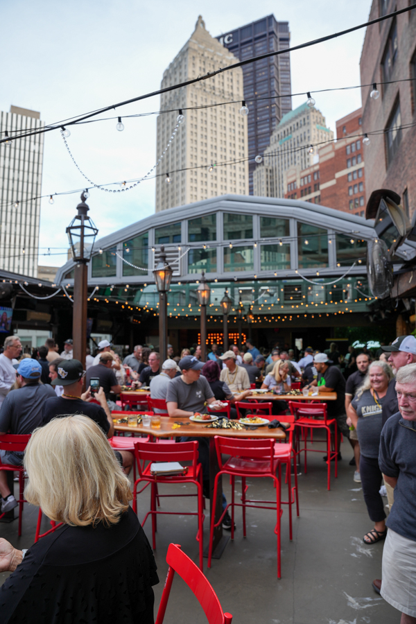 Crowded outdoor dining courtyard with red tables and chairs under a glass-roofed pavilion, string lights, and city skyscrapers in the background.