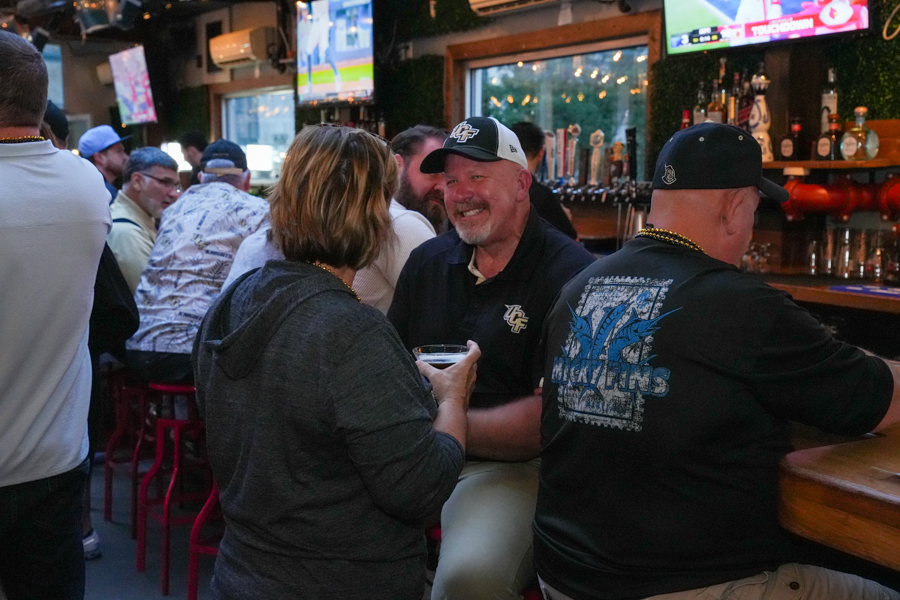 Group of people at a bar chatting and laughing, with taps and screens in the background, creating a lively social scene.