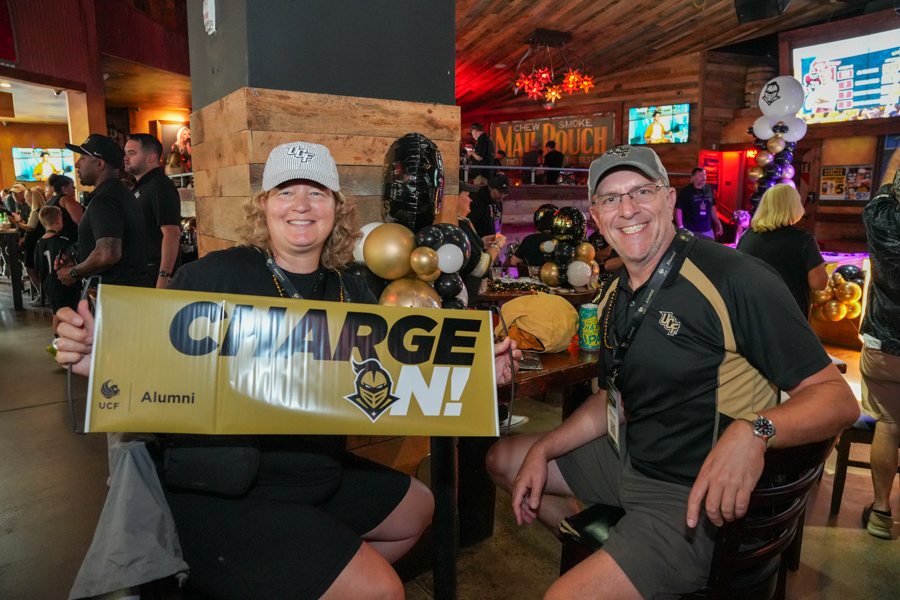 Two smiling people hold a gold 'CHARGE ON!' banner at a lively indoor alumni event with balloons in the background.