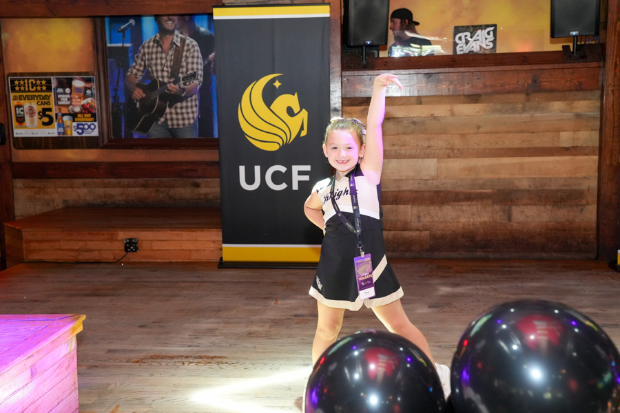 Young girl in a cheer outfit posing on a wooden stage with one arm raised, a UCF banner behind her and balloons in the foreground.