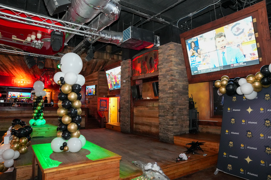 Balloon columns in black, white, and gold flank a wooden stage with green lighting at a bar venue.