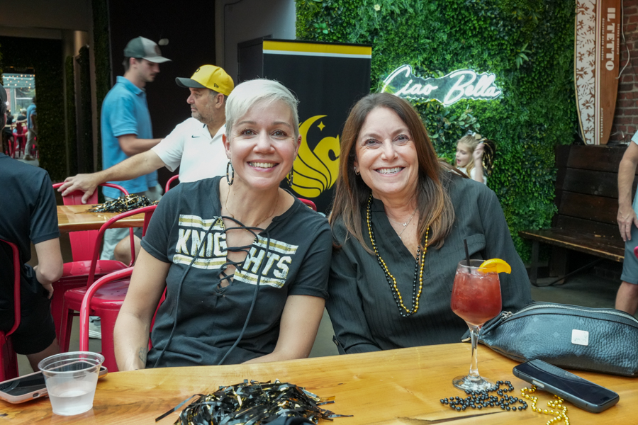 Two smiling women sit at a wooden table at a casual outdoor gathering, with drinks and decorations on the table.