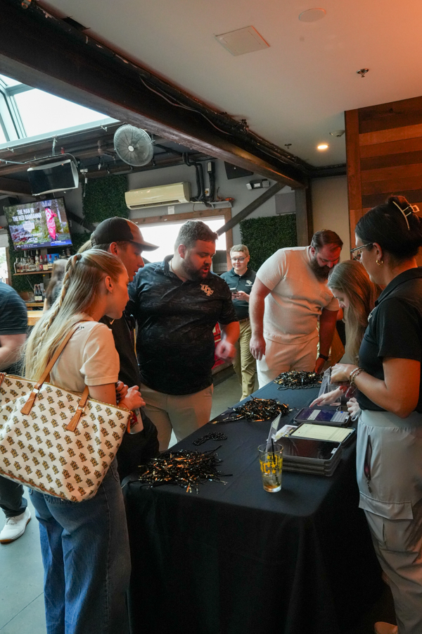 Group of people gathered around a table with black merchandise and a staff member assisting attendees at an indoor event venue.