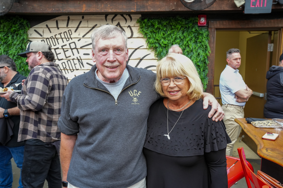 Two smiling older adults pose together, man with a gray quarter-zip and woman with blonde hair and glasses, arm around each other at a casual gathering.