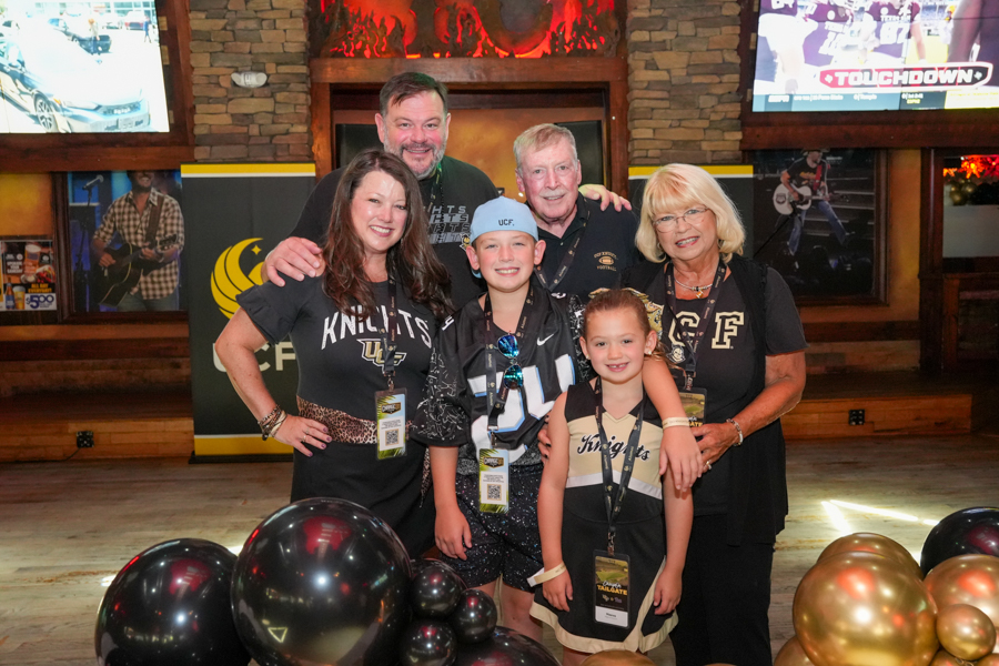 Six people posing for a group photo at a Knights sports event, with black and gold balloons in the foreground and TVs in the background.