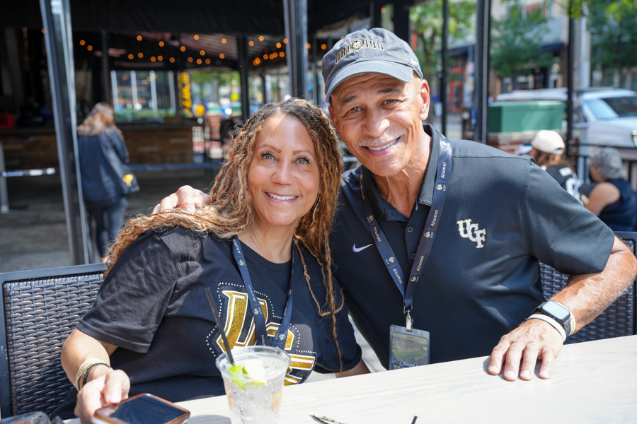 Two smiling adults sit at an outdoor cafe table with drinks, wearing lanyards and dark shirts.